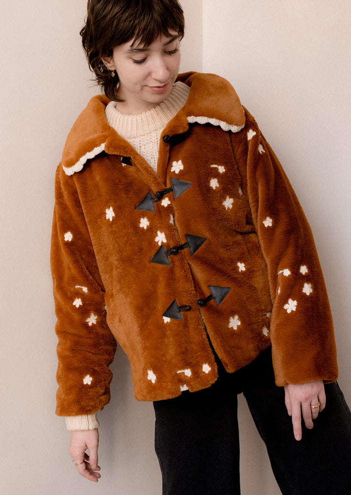 A woman wearing a caramel colored faux fur jacket with white floral embroidery and black toggle button front.