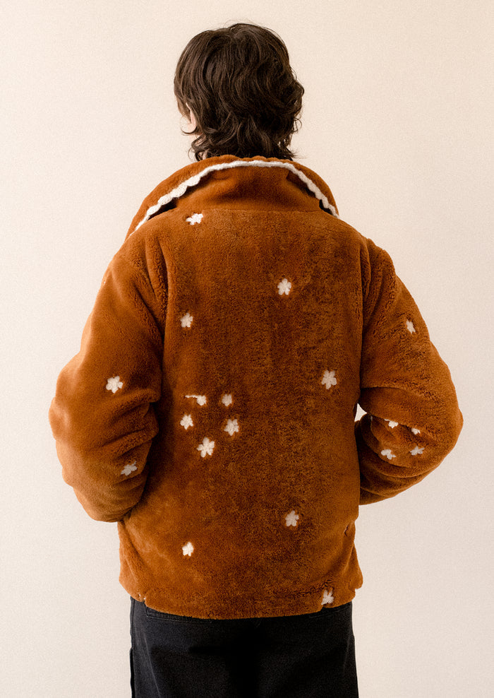 A woman wearing a caramel colored faux fur jacket with white floral embroidery and black toggle button front.