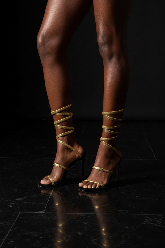 Black and gold spiral wrap stiletto sandals on a medium brown-skinned Black woman, dark stone floor, editorial luxury